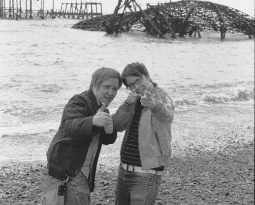 Two people posing in a desert-like setting with a dilapidated structure in the background.