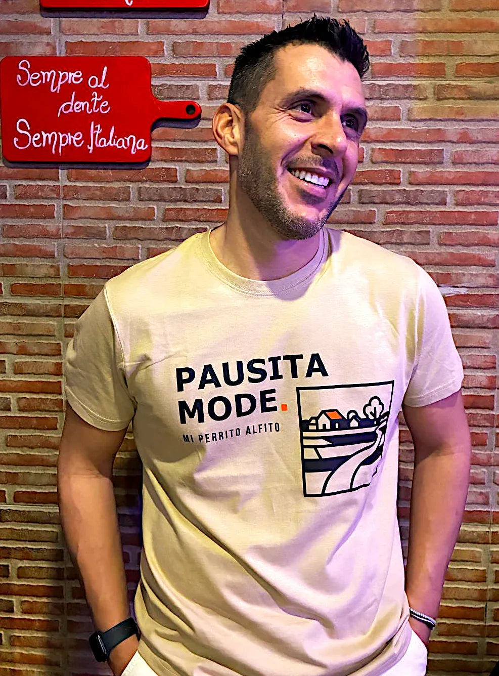 Man wearing a 'PAUSITA MODE' t-shirt against a brick wall with a red sign.